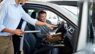 A man sitting in a car being instructed on how to use the vehicle by a woman with a tablet